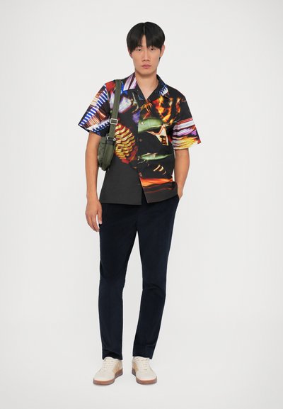 Maglietta a maniche corte con vivaci motivi astratti, base nera, abbinata a pantaloni blu navy e sneakers chiare. Borsa a tracolla in verde oliva.