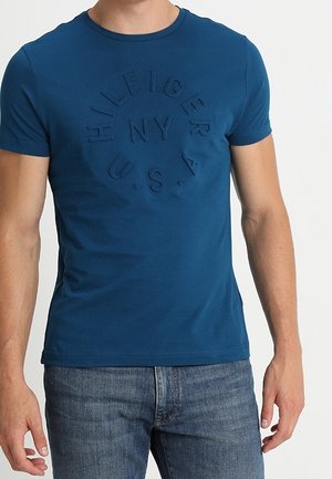 Uomo che indossa una maglietta blu aderente con testo circolare in rilievo "HILFIGER NY U.S.A." e jeans blu su uno sfondo semplice.