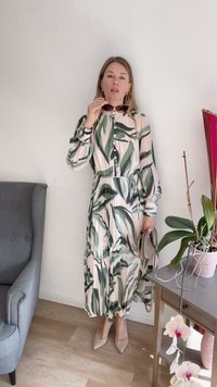Vestido verde com padrão de folhas, com mangas longas e botões, cintura ajustada e tecido fluido. Usado com sapatos de salto bejes e segurando óculos de sol.