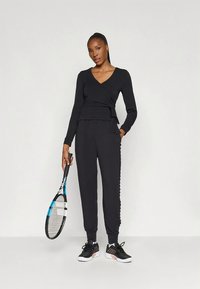 Γυναίκα με μαύρη αθλητική στολή που κρατάει ένα ρακέτα τένις, στέκεται μπροστά σε έναν απλό ανοιχτό φόντο.