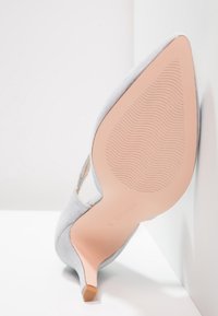 Grå mockahögklacksko med spetsig tå, visar en texturerad rosa sula och subtila utskärningsdetaljer på sidorna.