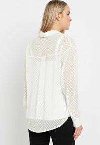 Weißes transparentes Bluse mit Fischgrätenmuster, langen Ärmeln, Kragen und abgerundetem Saum. Weicher, leichter Stoff.