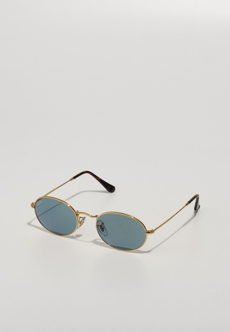 Óculos de sol com armação dourada em metal, lentes azuis, formato oval e ponteiras castanhas. As características notáveis incluem hastes finas e almofadas nasais transparentes.