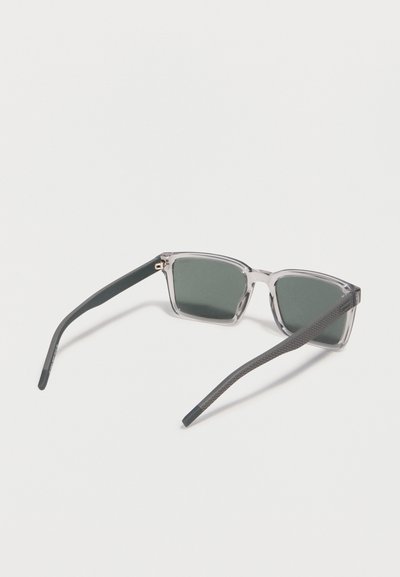 Lunettes de soleil rectangulaires avec montures gris transparent et verres vert foncé reposant ouvertes sur une surface blanche.