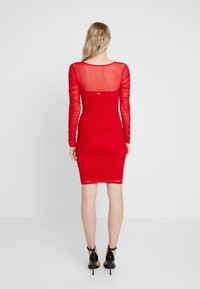 Robe rouge avec manches longues en voile transparent, texture froncée sur toute la surface, silhouette ajustée et ourlet à hauteur des genoux. Talons noirs portés avec la robe.