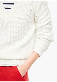 Pull en tricot blanc avec un col en V, motif texturé et poignets côtelés. Associé à un pantalon rouge, soulignant un look décontracté.