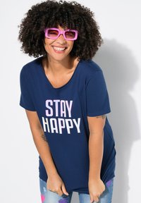Námořnicky modré tričko s nápisem "STAY HAPPY" v růžové a bílé. Modelka má na sobě oversized růžové sluneční brýle. Ležérní, krátký rukáv a kulatý výstřih.