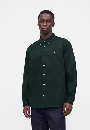 Uomo che indossa una camicia verde scuro con bottoni e un piccolo logo bianco sul petto e jeans blu scuro, in piedi davanti a uno sfondo semplice.