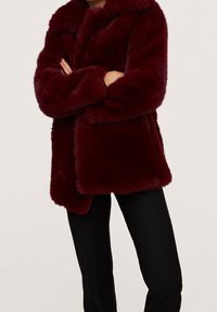 Persona que lleva un abrigo de piel sintética de color rojo oscuro con los brazos cruzados, combinado con pantalones negros, de pie frente a un fondo claro y uniforme.