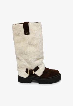 Bota de ante marrón con caña de piel sintética blanca y esponjosa, suela de goma negra y un detalle de hebilla lateral. Diseñada para uso en invierno.