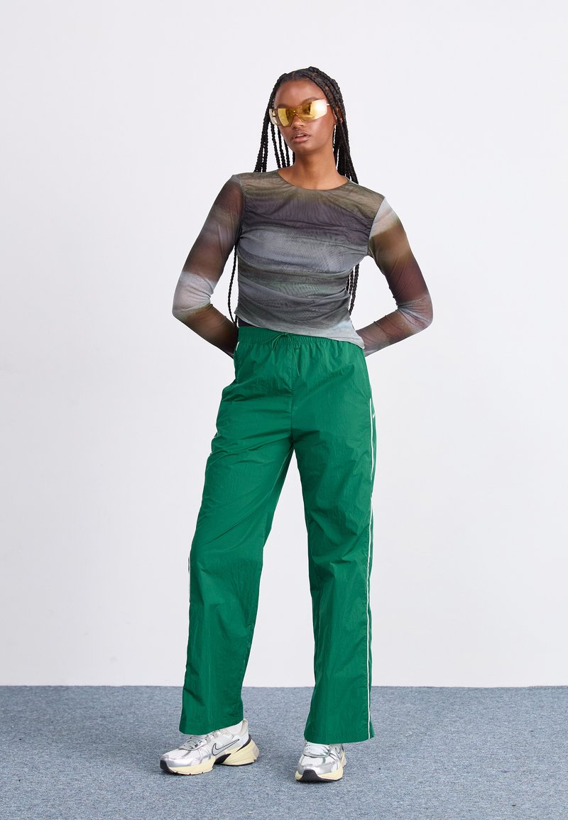 Transparante blouse met lange mouwen met draaiende bruine en grijze patronen; groene sportbroek met witte zijstreep; zilveren sneakers; gele zonnebril.
