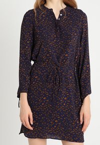 Granatowa sukienka z lekkiego materiału z kwiatowym wzorem w odcieniach brązu, z guzikiem przy dekolcie, długim rękawem i regulowanym paskiem w talii.
