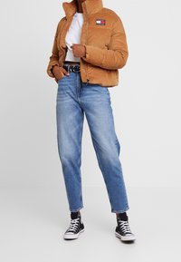 Brun kordflasket pufferjacka med hög krage och en liten logopatch, kombinerad med ljusblå raka jeans och svarta Converse-sneakers.