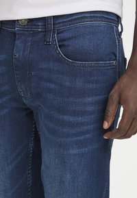 Temno modri jeans iz denima z gladko teksturo, dizajn s petimi žepi in subtilnimi gubami ob stegnu. Kovinski gumb za zapiranje na pasu.