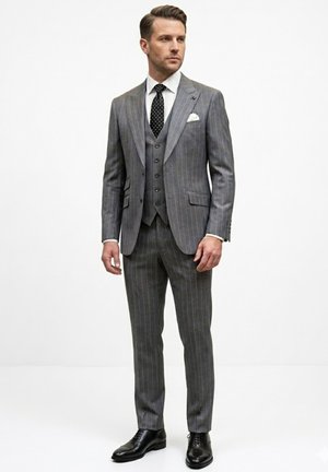 Hombre con traje de tres piezas de rayas grises, camisa blanca, corbata negra con lunares blancos, pañuelo blanco en el bolsillo y zapatos de vestir negros, de pie erguido.