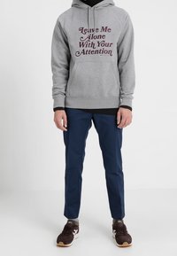 Grå huvtröja med mörkröd text, marinblå byxor och mörkbruna sneakers. Huvtröjan har dragsko och ribbade muddar.