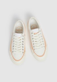 Zapatillas de lona blancas con acentos melocotón, punta redonda y diseño de cordones. Presentan ojales metálicos y suela de goma texturizada.