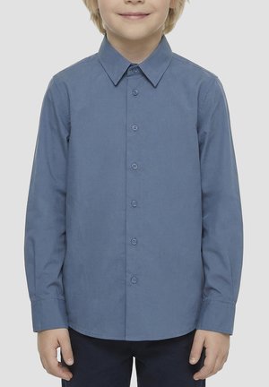 Bambino che indossa una camicia blu a maniche lunghe con bottoni e colletto, in piedi con le braccia rilassate su uno sfondo semplice.