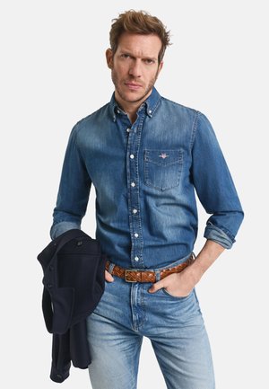 Denim button-up shirt in light blue, featuring a chest pocket. Pairing with light blue jeans and a brown woven belt. Black jacket held in left hand.