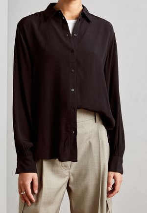 Osoba nosící volnou černou košili na knoflíky přes bílý top, doplněná světle béžovými kalhotami s vysokým pasem, sklady a kapsami.