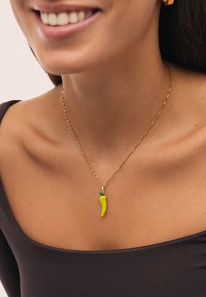 Collar de cadena de oro que presenta un colgante en forma de chile amarillo con un acento verde. El modelo lleva una blusa marrón oscuro, destacando el escote y el colgante.