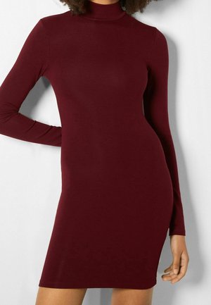 Woman wearing a fitted, long-sleeve, burgundy turtleneck dress, standing with one arm bent and the other down by her side.
