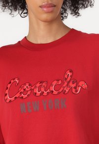 Rotes Baumwoll-T-Shirt mit einem gestickten "Coach"-Logo im Schlangendesign, mit glatter Textur und einem abgerundeten Ausschnitt.