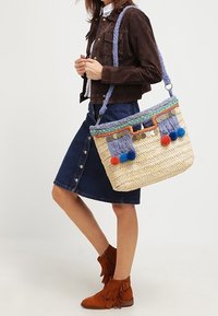 Woven straw bag with blue and orange pom-poms, decorative tassels, and a braided strap. Features a colorful trim and metallic accents.