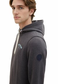 Felpa grigia con cappuccio realizzata in tessuto morbido, presenta un cordino, un logo azzurro chiaro e una toppa azzurra navy sul maniche sinistro.