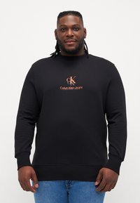 Crna dukserica dugih rukava, s rebrastim manžetama i donjim rubom. Na prednjoj strani u središtu sadrži logotip "Calvin Klein Jeans" u narandžastoj boji. Materijal je mješavina pamuka.