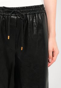 Gros plan sur un pantalon noir en faux cuir avec une taille élastique et des cordons noirs à bouts dorés, porté avec un haut blanc.