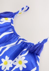 Tirante azul de vestido con tela fruncida, estampado de margaritas blancas y amarillas y detalle de borla azul sobre fondo claro.