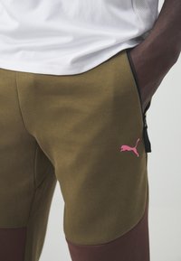 Pantalon de survêtement vert olive avec un panneau marron foncé, arborant un logo Puma rose, des poches zippées sur les côt és et une texture lisse et douce.