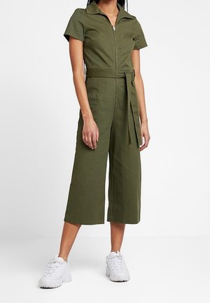 Jumpsuit verde oliva con maniche corte, zip frontale, vita con cintura e gambe larghe e cropped, abbinato a sneakers bianche. Tessuto di cotone.