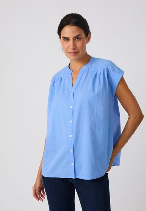 Vrouw die een losse lichtblauwe mouwloze blouse met knopen en donkere spijkerbroek draagt, staand met één hand achter haar rug tegen een effen achtergrond.