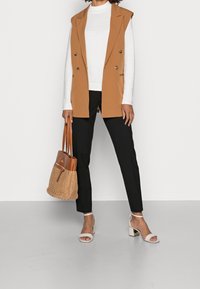 Žena v béžové bezrukávové blazer, bílém žebrovaném tričku s dlouhým rukávem, černých kalhotách a bílých sandálech na blokovém podpatku, držící béžovou pletenou kabelku.