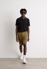 Polo nero con dettagli bianchi, pantaloni cargo verde oliva e sneakers bianchi. Il modello indossa anche occhiali da sole neri. Sfondo minimalista.