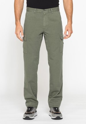 Uomo che indossa pantaloni cargo verde oliva con tasche applicate e sneakers grigio nere, in piedi davanti a uno sfondo semplice.