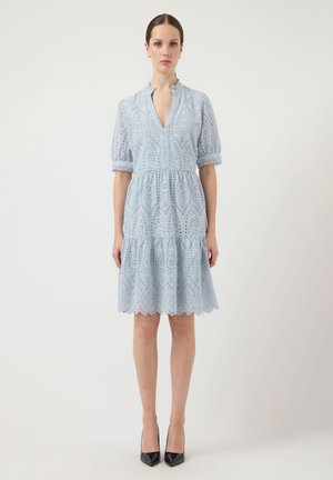 Moteris stovi šviesiai mėlynoje su akelėmis suknele su trumpomis pūstomis rankovėmis, raukšlėtu apykakle ir nelygiu apdailos kraštu, dėvi juodus smailius aukštakulnius.