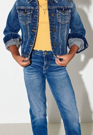 Denim-takki, jossa on kaksi etutaskua, sininen väri ja napitus, yhdistettynä keltaiseen ribattuun toppiin ja tummansinisiin farkkuihin.