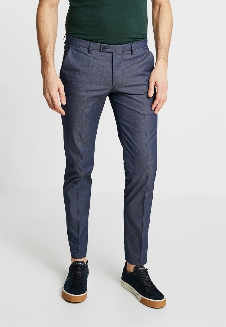 Homme portant un pantalon de costume bleu foncé ajusté et des baskets noires à semelles blanches, debout devant un fond clair uni.