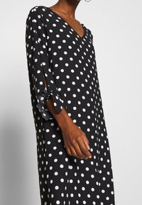 Musta mekko valkoisilla pilkuilla, V-kaula-aukko, puolipitkät hihat halkioilla ja sidotuilla naruilla, jota pukeutuu keskiruskea iho.