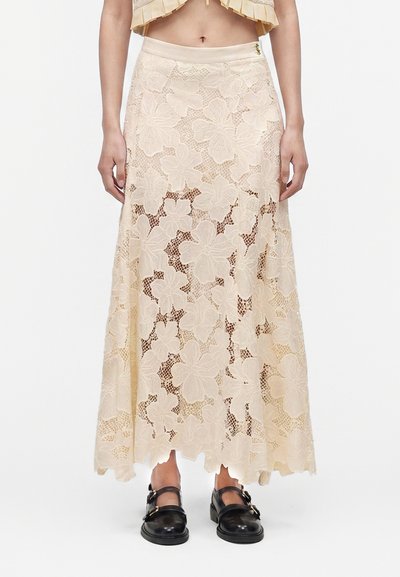 Jupe maxi en dentelle florale beige portée avec un top court et des chaussures noires style Mary Jane, montrant de la taille aux pieds sur fond blanc.