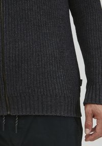 Strikkegenser i mørk sjøgrønn med teksturert mønster, glidelås foran, ribbestrikkede mansjetter og nederkant, og en liten logoetikett på siden.