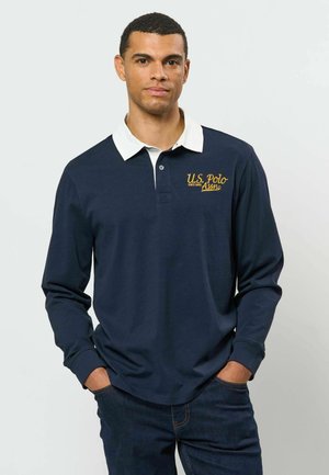 Homme portant un polo bleu marine à manches longues avec un col blanc et un logo jaune U.S. Polo Assn., mains dans les poches, debout devant un fond uni.