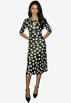 Schwarz-weißes Midi-Kleid mit Punkten, kurzen Puffärmeln, einem Keyhole-Ausschnitt und taillierter Silhouette. Kombiniert mit schwarzen High Heels.