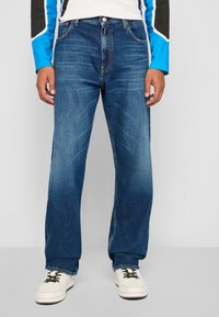 Jean en denim bleu foncé avec une coupe décontractée, comportant un design à cinq poches, des décolorations subtiles et des détails cousus. Associés à des baskets blanches.
