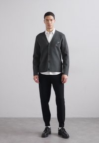 Cardigan grigio scuro con scollatura a V, cinque bottoni e un emblema ricamato. Abbinato a una camicia bianca, pantaloni neri e scarpe in pelle.