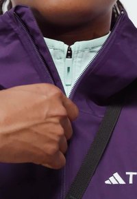 Ljubičasta jakna s unutarnjom podstavom u mint zelenoj boji, koja ima prednji zip i crnu traku. Istaknut Adidas logo na donjoj strani.