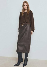 Top marrón de manga larga, falda de cuero marrón de talle alto y botas negras de tacón con punta afilada. Tela texturizada y un cinturón con hebilla dorada como detalle.